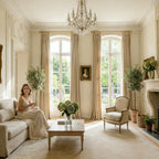 Lifestyle view of a classic French-style living room featuring tied-back beige Adrian cashmere blackout curtains over arched windows, with a woman sitting on the sofa.