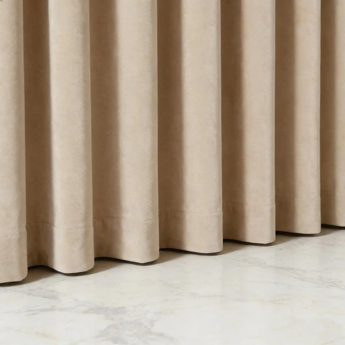 Low-angle close-up detail of the neat bottom hem of beige Adrian extra long and wide cashmere blackout curtains resting on a polished marble floor.
