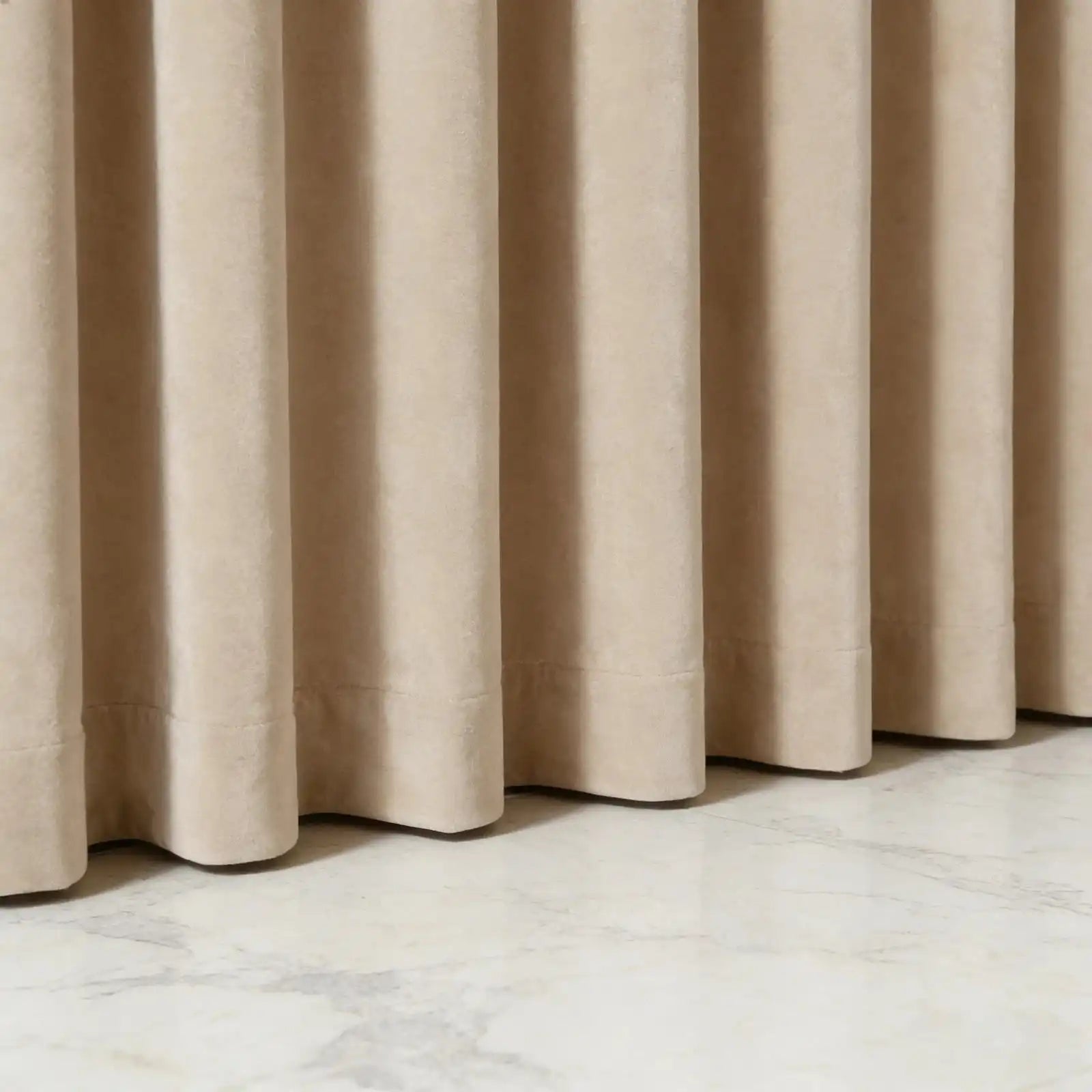 Low-angle close-up detail of the neat bottom hem of beige Adrian extra long and wide cashmere blackout curtains resting on a polished marble floor.