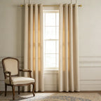 Pair of Adrian beige grommet-top cashmere blackout curtains hanging on a brass rod in a classic living room with an armchair.