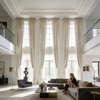 Three pairs of custom Adrian extra long and wide beige cashmere blackout curtains installed in a luxury double-height modern living room with high ceilings.