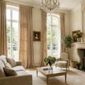Pair of custom beige Adrian cashmere blackout curtains tied back on tall arched windows in a classic French-style living room with a fireplace.