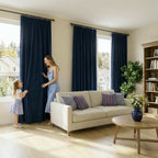 A living room scene capturing the mother and daughter playfully pulling back a deep sea blue curtain panel, with a cream sofa, a round wooden coffee table, and a tall wooden bookshelf visible.