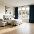 A cozy bedroom setting displaying a bed with a light gray tufted headboard, an armchair with a blue plaid pillow on the left, and deep sea blue curtains dressing the far window under soft sunlight.