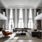 A wide-angle shot of a grand, luxurious living room with high coffered ceilings, showcasing six towering gray curtain panels framing massive windows behind a large gray sectional sofa and a modern orange accent chair.