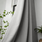 A close-up shot of the gray curtain fabric being pulled back beside a white window frame, flanked by a slender green houseplant on the left.