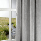 A close-up view of a light gray curtain panel hanging neatly next to a white-framed window that looks out onto a vibrant green field with a winding stream.