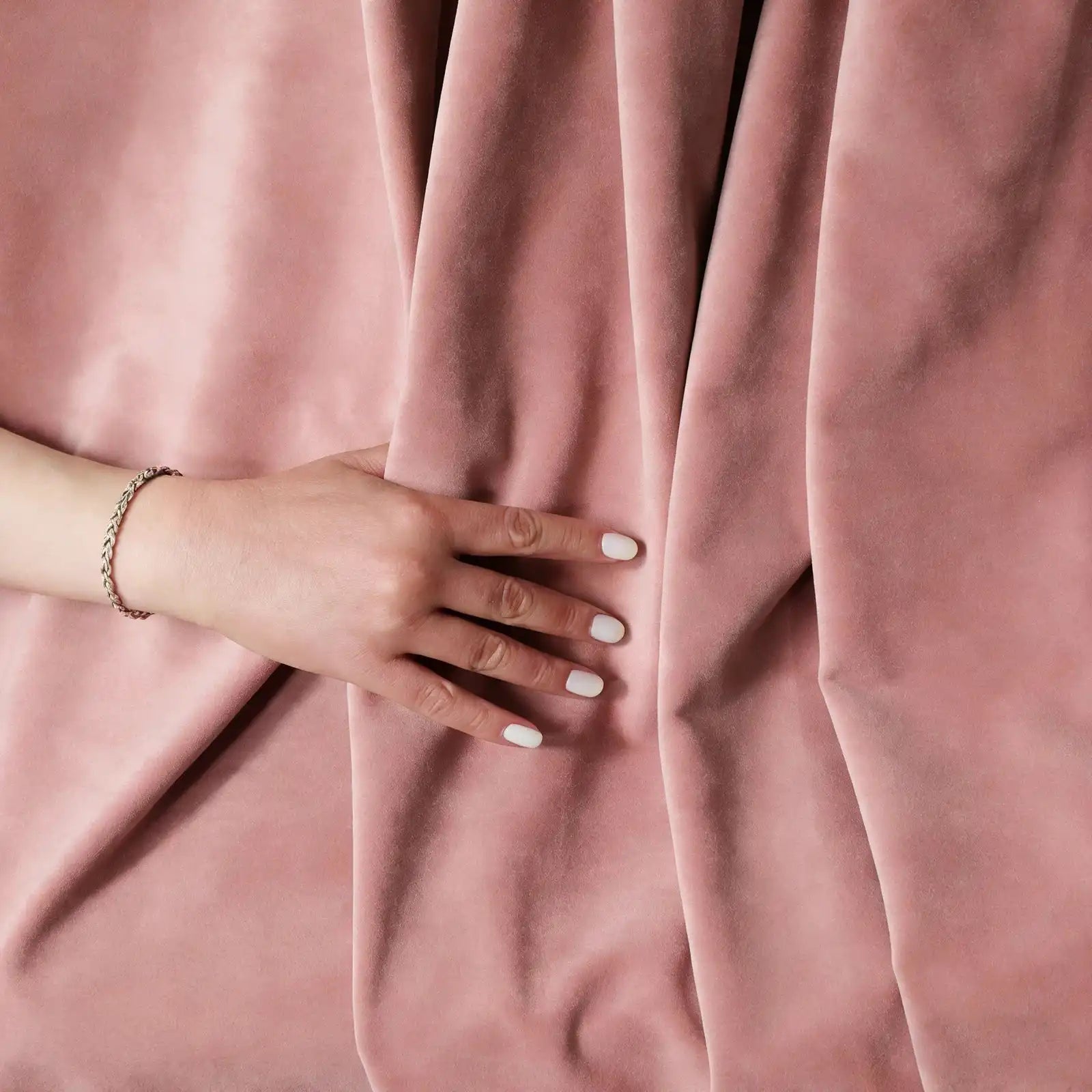 A top-down view of a hand with a silver bracelet gently resting on the plush, folded pink curtain fabric, emphasizing its soft, touchable texture.