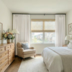 A straight-on, frontal shot of the bright bedroom setup, highlighting the crisp, clean appearance of the off-white curtains framing the large window, the cozy reading corner, and the edge of the bed.