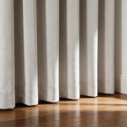 A close-up shot focusing on the bottom hem of the off-white curtains as they pool gracefully onto a polished dark wood floor, illuminated by a warm beam of sunlight.