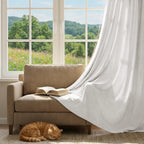 A cozy, sunlit lifestyle scene featuring an off-white curtain elegantly draped over a beige armchair with an open book. A ginger cat is sleeping on the rug below, next to a large window overlooking a scenic green hill landscape.