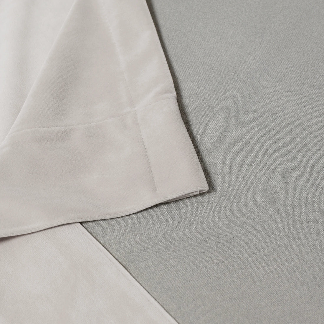 A structural close-up shot detailing the neat, professional corner stitching and hem of the off-white curtain fabric resting flat against a light gray textured surface.