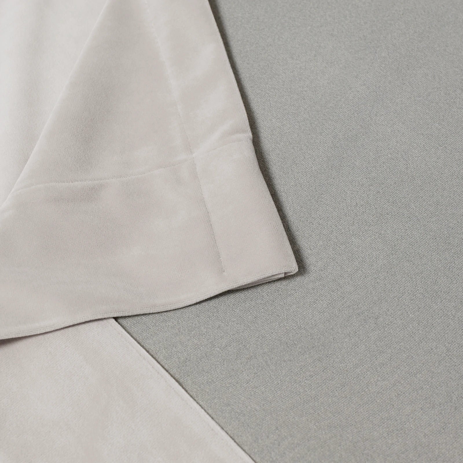 A structural close-up shot detailing the neat, professional corner stitching and hem of the off-white curtain fabric resting flat against a light gray textured surface.