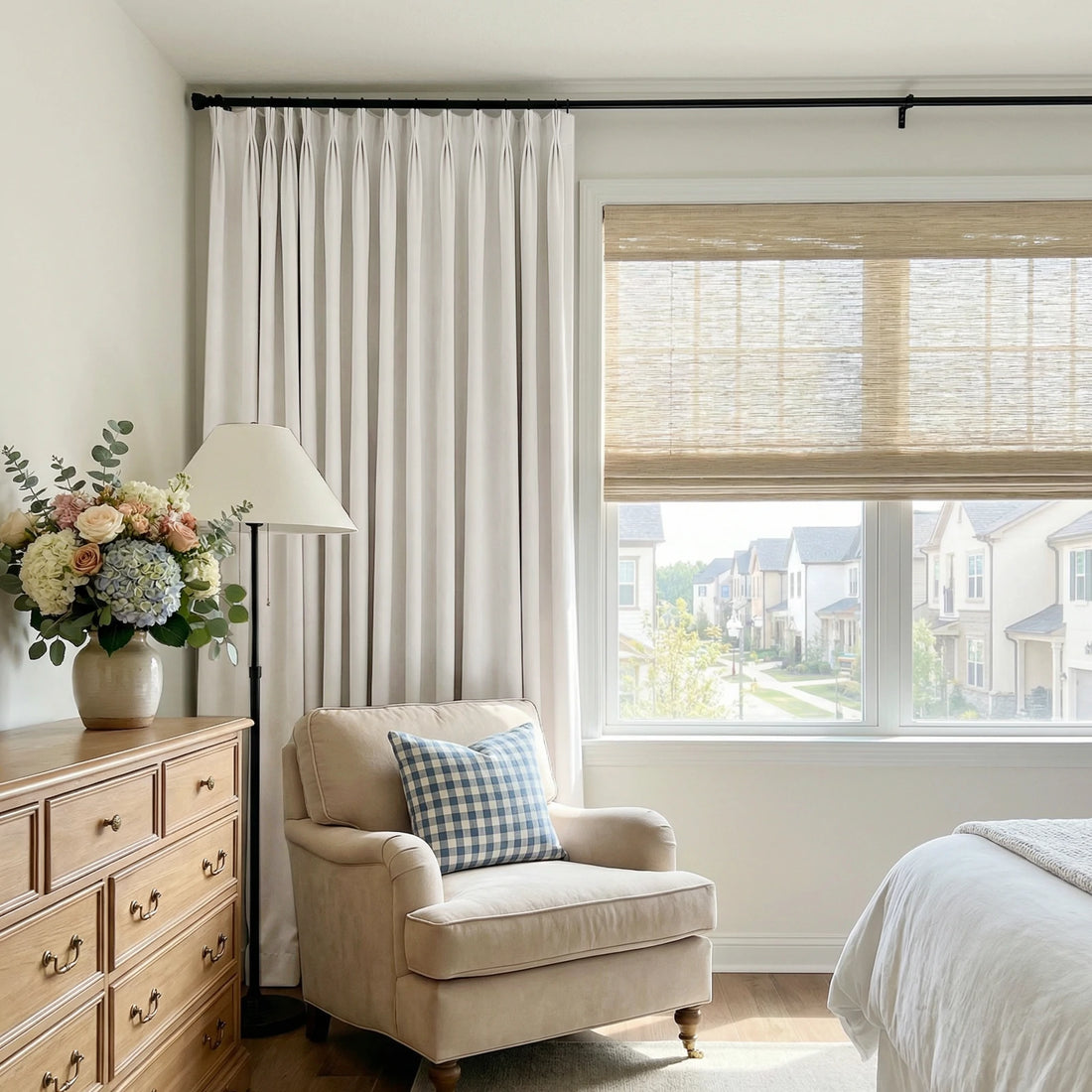 A bedroom view from the left side, showing a wooden dresser topped with a colorful flower bouquet, a beige armchair with a blue checkered pillow, and a single panel of off-white pleated curtains hanging next to a window with a woven bamboo blind.