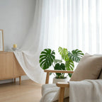 Bright living room with white semi-sheer curtains, a wooden sideboard, a potted monstera plant, and a beige armchair with a textured throw blanket