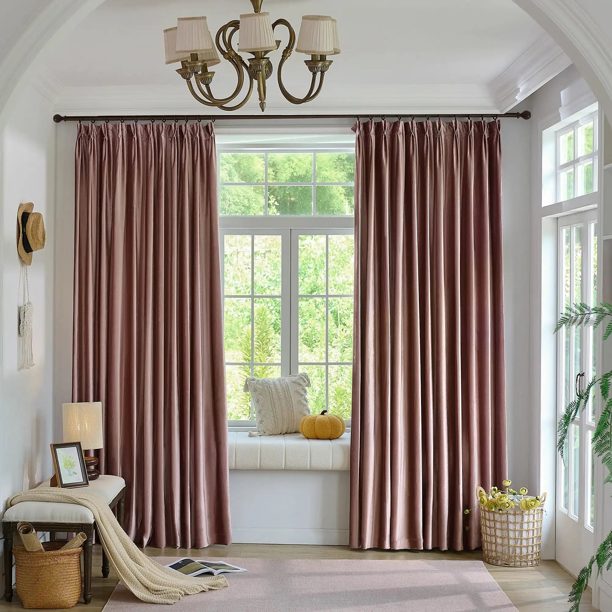 Pink velvet curtains hanging in front of a large window with greenery outside, decorated with a small yellow pouf and a cozy window seat