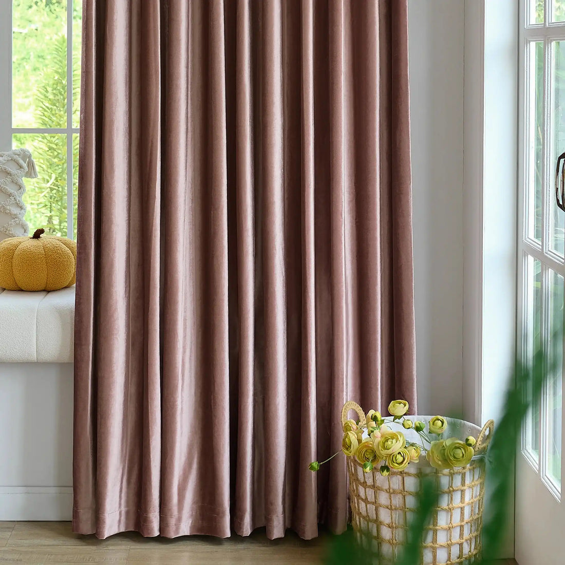 Pink velvet curtains hanging in front of a large window with greenery outside, decorated with a small yellow pouf and a cozy window seat
