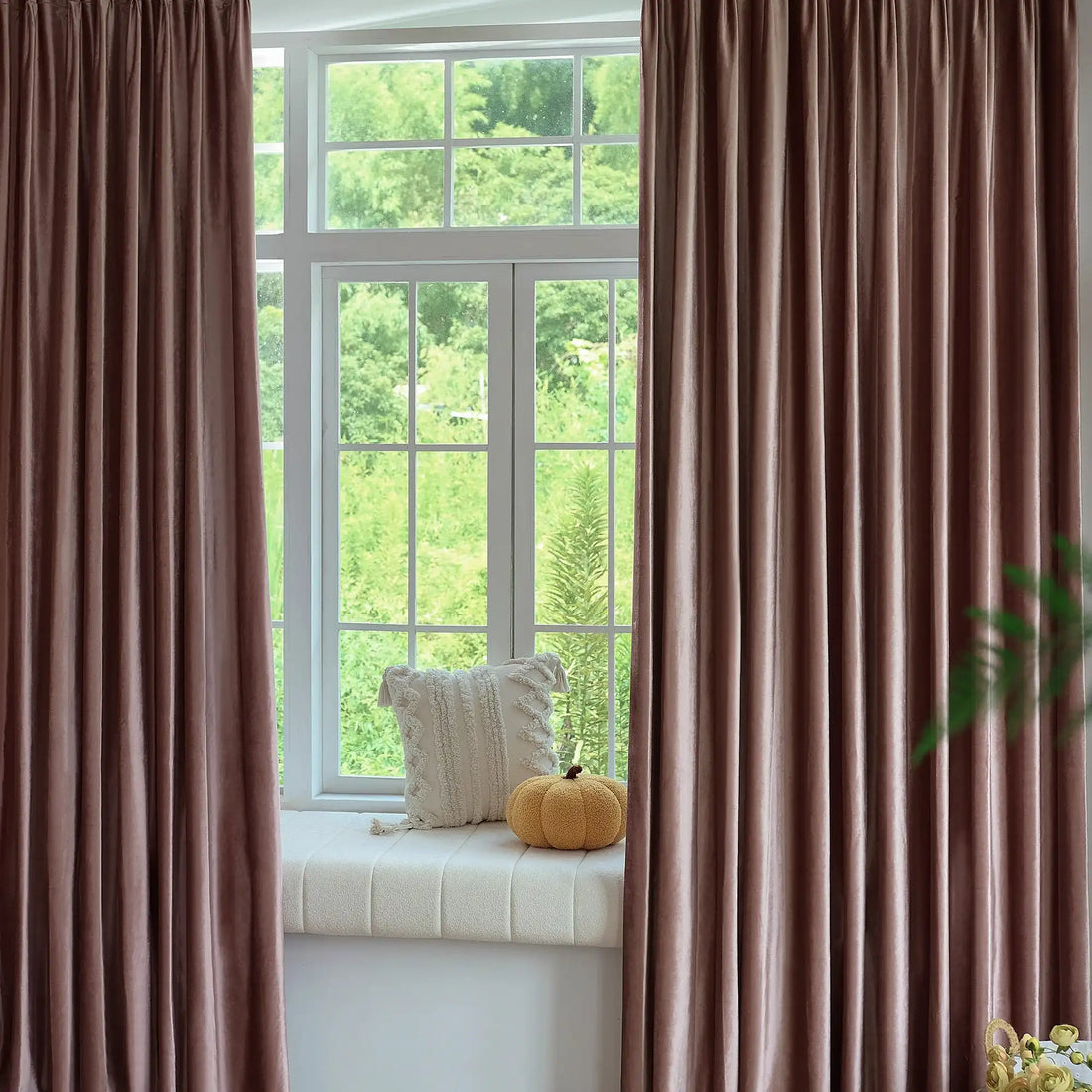 Pink velvet curtains hanging in front of a large window with greenery outside, decorated with a small yellow pouf and a cozy window seat
