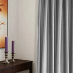 Gray velvet curtains with textured fabric hanging on a wooden rod, in a minimalist room with white walls and a large window.