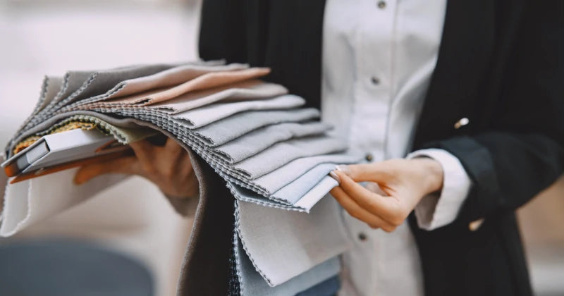 A person in a black blazer is holding and examining a fan of neutral-colored fabric swatches?aideal for choosing materials for custom Caleb Cross-Weave Privacy Curtains by ArgaHome. Only the individual?¡¥s torso and hands are shown.