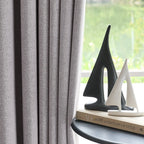 ArgaHome Orion Linen Look 100% Blackout Curtains in gray hang beside a window, while two modern sailboat sculptures?aone black and one white?aare displayed on top of a stack of books on a small round table. Sunlight streams in through the window.