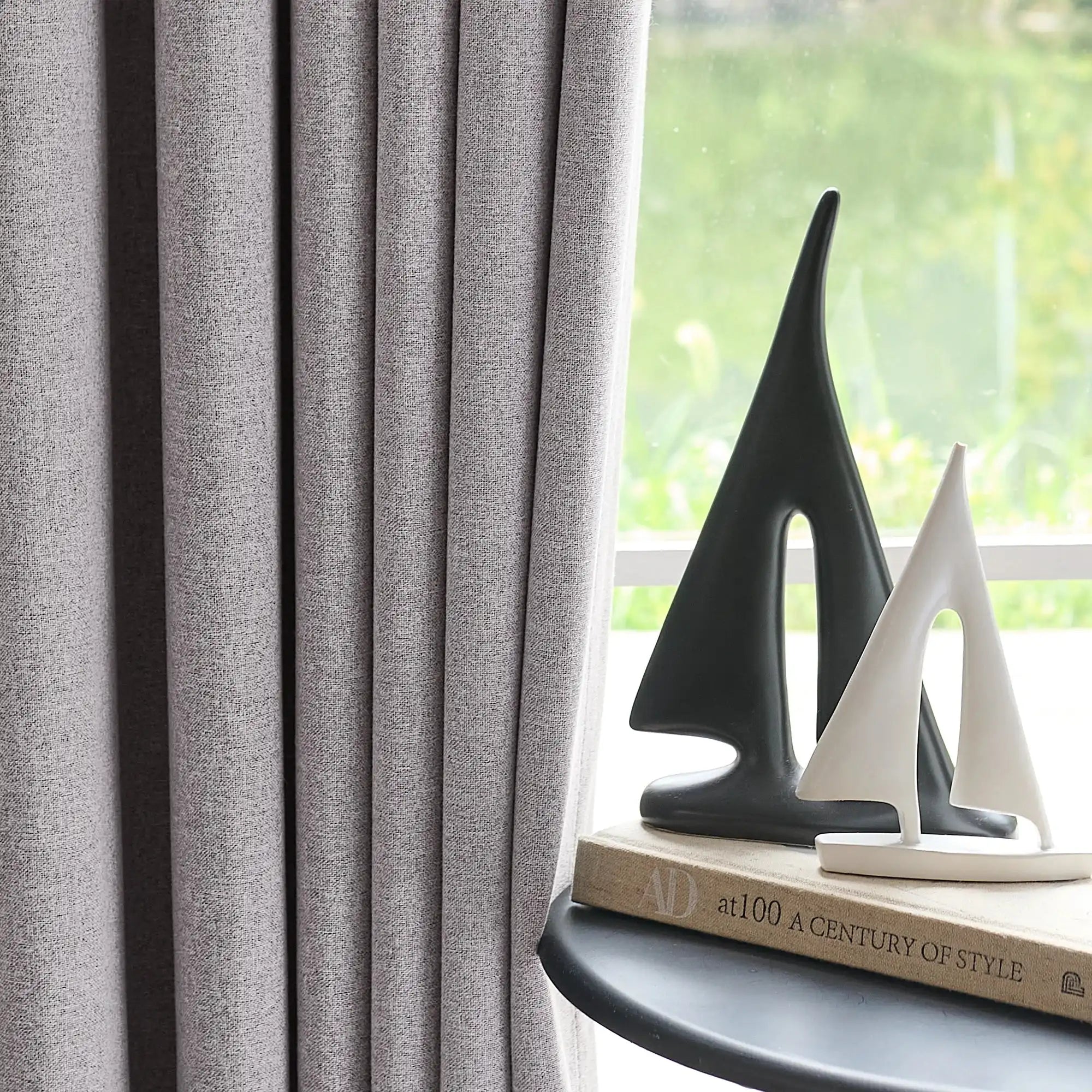 ArgaHome Orion Linen Look 100% Blackout Curtains in gray hang beside a window, while two modern sailboat sculptures?aone black and one white?aare displayed on top of a stack of books on a small round table. Sunlight streams in through the window.