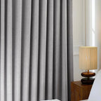 An ArgaHome Orion Linen Look 100% Blackout Curtain is closed beside a bed, with a beige table lamp featuring a woven shade placed on a wooden nightstand in the softly lit room.