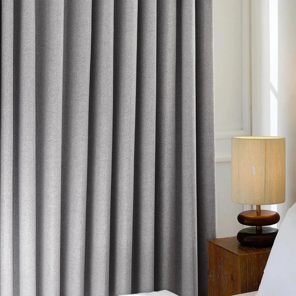 An ArgaHome Orion Linen Look 100% Blackout Curtain is closed beside a bed, with a beige table lamp featuring a woven shade placed on a wooden nightstand in the softly lit room.