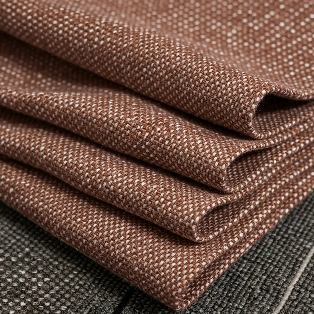 A macro photograph of the caramel fabric neatly folded into precise, crisp diagonal layers. The shot highlights the substantial weight and textured weave of the textile as it rests against a dark gray mottled surface.