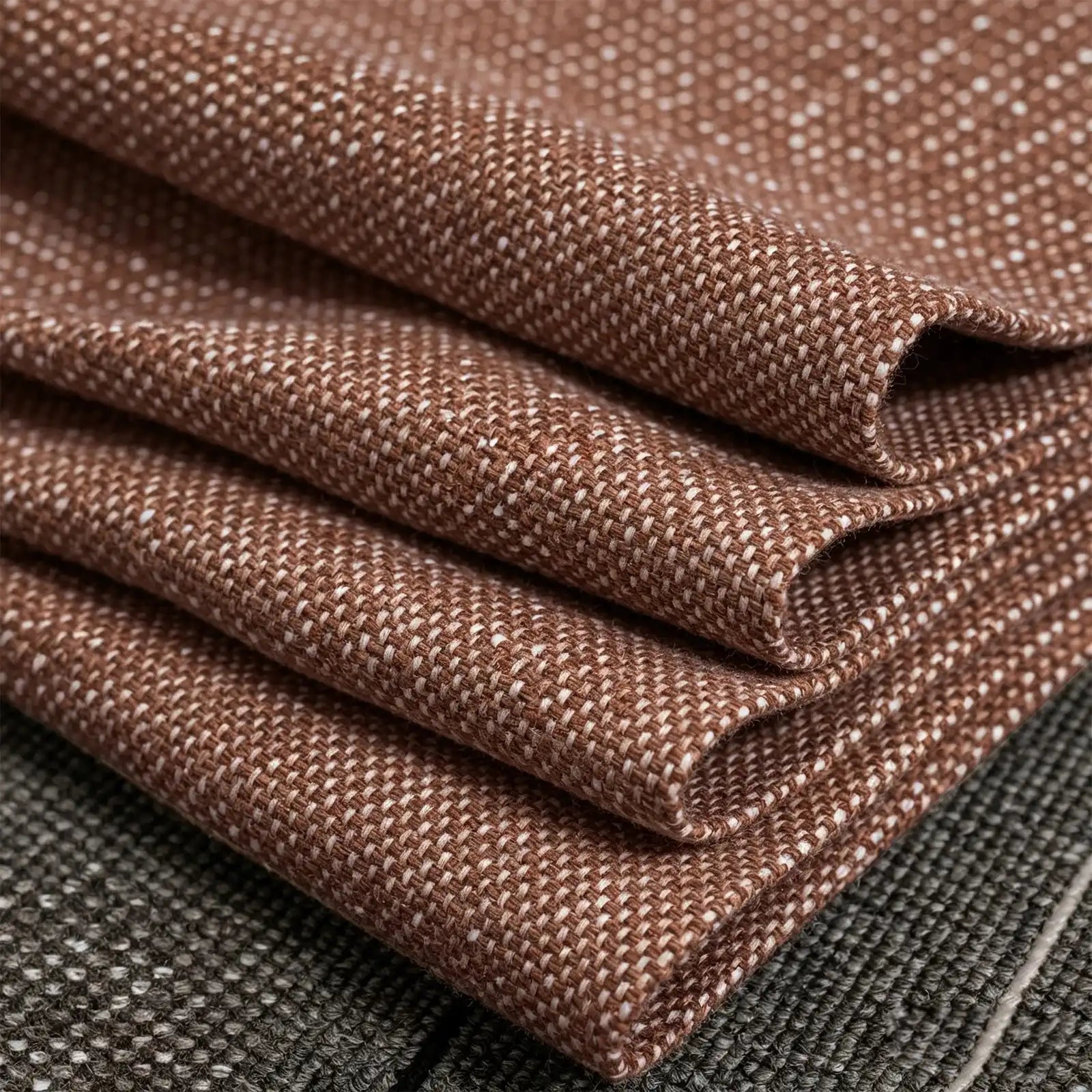 A macro photograph of the caramel fabric neatly folded into precise, crisp diagonal layers. The shot highlights the substantial weight and textured weave of the textile as it rests against a dark gray mottled surface.