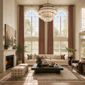A dramatic, wide-angle view of a grand living room with soaring ceilings and double-height arched windows. Long, elegant caramel curtain panels frame the large windows. The space features a classic fireplace, a long off-white sofa, a dark rustic wood coffee table, and plaid upholstered stools.