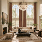 A dramatic, wide-angle view of a grand living room with soaring ceilings and double-height arched windows. Long, elegant caramel curtain panels frame the large windows. The space features a classic fireplace, a long off-white sofa, a dark rustic wood coffee table, and plaid upholstered stools.