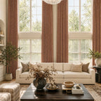 A closer, front-facing view of the high-ceilinged living room, centering on the cream-colored sofa styled with decorative throw pillows. In the foreground, a dark wooden coffee table holds a prominent ceramic vase with dried branches, framed by the long, dramatic columns of caramel drapery.