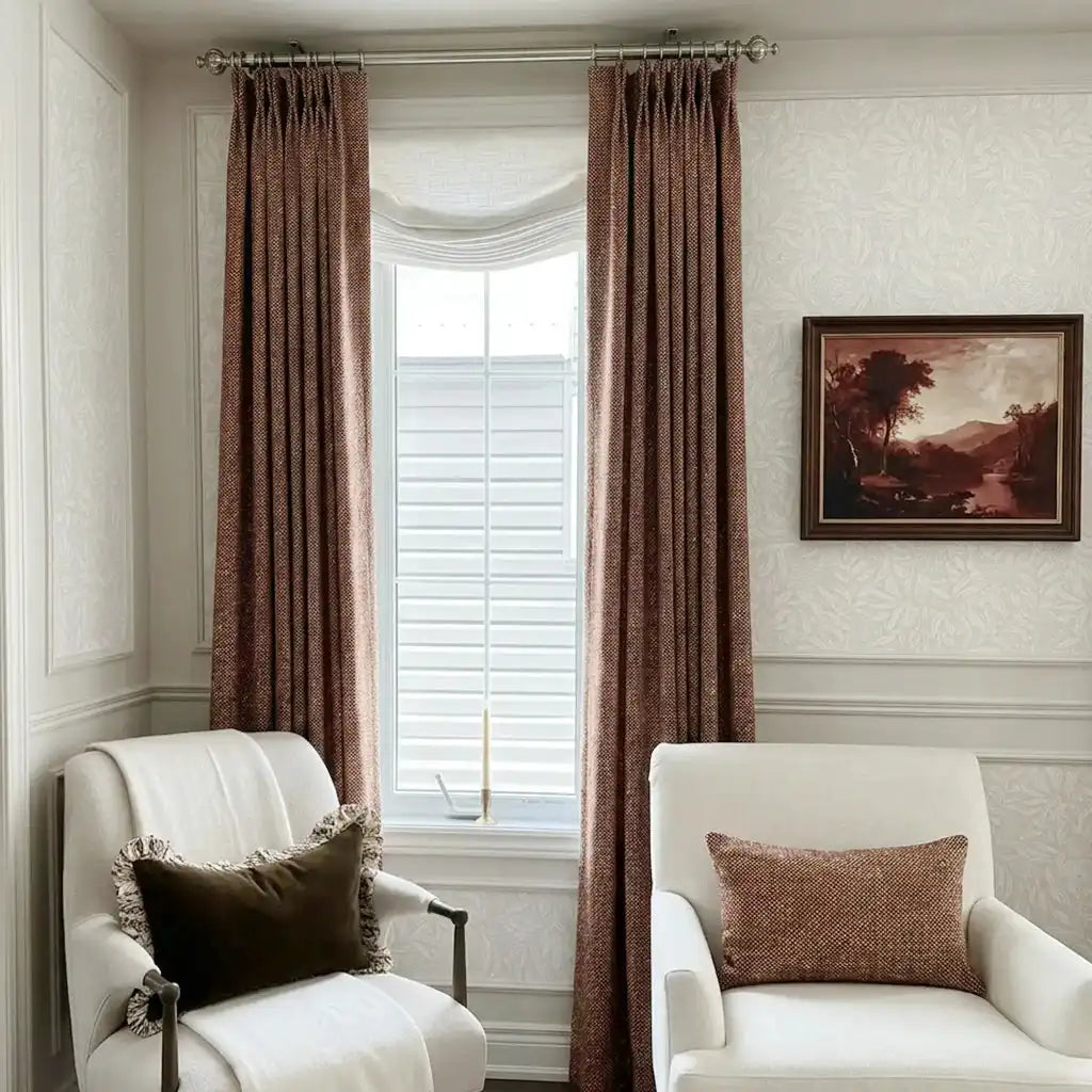 A beautifully styled traditional room corner featuring a window dressed with long, pleated caramel-colored curtains and a soft white Roman shade. Two comfortable white armchairs flank the window?aone with a dark brown fringed pillow and the other with a matching caramel patterned pillow?abeneath a classic framed landscape painting.