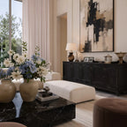 An interior view of the luxury living room setup, focusing on a black marble coffee table adorned with fresh white and blue flowers, a cream ottoman, and a dark carved wooden credenza beneath a large abstract painting.