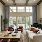 A beautifully centered, wide-angle view of the luxurious living room, displaying the harmonious layout of the live-edge wooden coffee table topped with books, flanked by red leather chairs and the curved cream sofa beneath the towering windows and gray drapery.