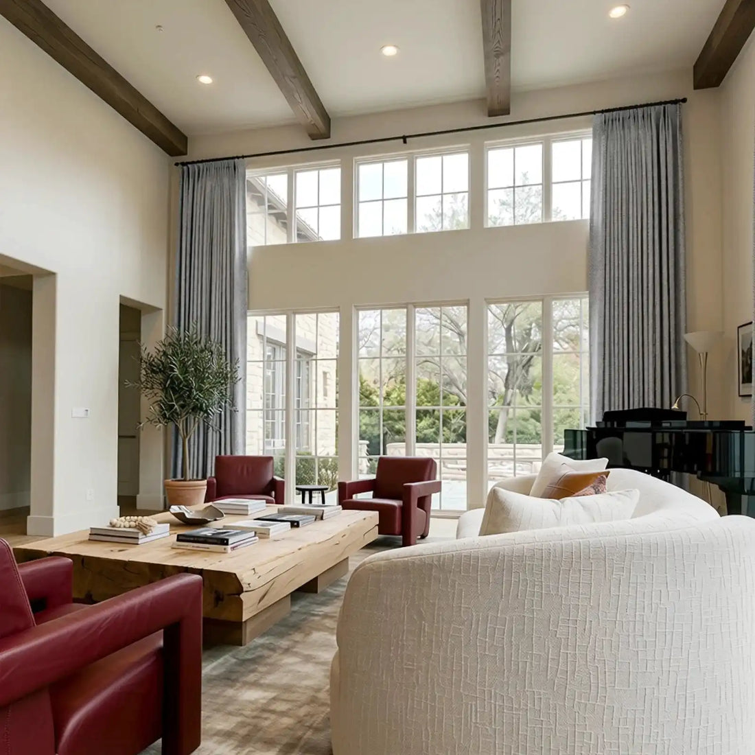 A grand, high-ceilinged luxury living room with exposed dark wood ceiling beams and massive multi-pane windows framed by long, elegant gray pleated curtains. The sophisticated space features a modern cream sofa, deep red leather armchairs, a large rustic live-edge wooden coffee table, and a glossy black grand piano.
