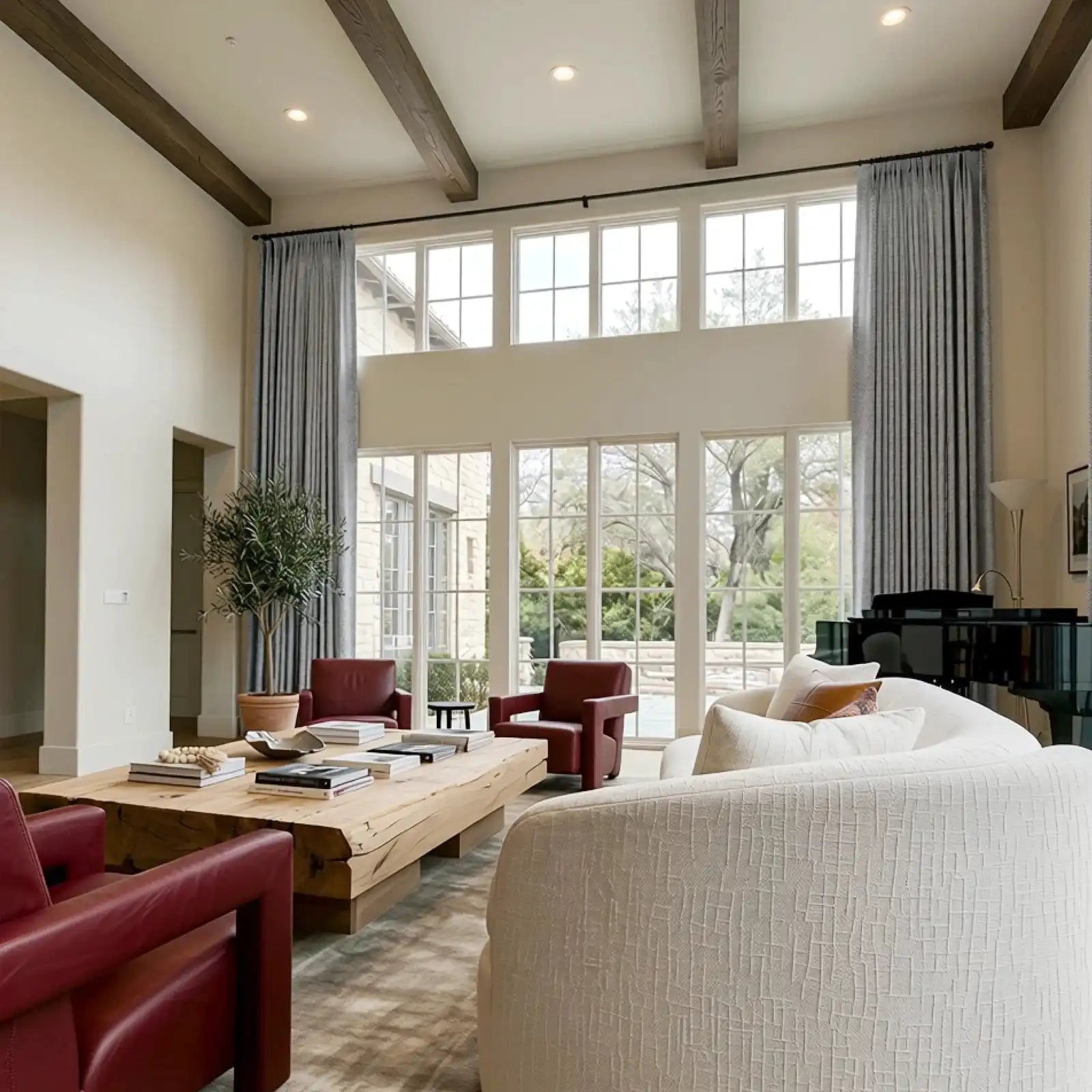 A grand, high-ceilinged luxury living room with exposed dark wood ceiling beams and massive multi-pane windows framed by long, elegant gray pleated curtains. The sophisticated space features a modern cream sofa, deep red leather armchairs, a large rustic live-edge wooden coffee table, and a glossy black grand piano.