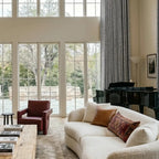 A closer interior shot focusing on the plush, curved cream sofa adorned with a smooth brown leather pillow and a patterned lumbar cushion. In the background, a stately black grand piano stands beside tall, meticulously pleated gray curtains hanging from a minimalist black rod.