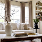 A close-up interior view focusing on a white marble coffee table with decorative books and a tall ceramic vase holding bare branches. In the background, a cream-colored sofa with striped pillows sits before a bay window dressed in elegant, grayish-brown pleated curtains.
