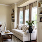 An angled perspective of the living room showing the right side of the cream sofa. A black pedestal side table holds a lush green plant in a dark ceramic jar, positioned next to a built-in white wall unit with an arched display shelf.