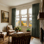 A warm, traditional sitting room featuring three classic wooden-frame armchairs upholstered in a vintage floral and plaid pattern. A central wooden coffee table holds a vase of flowers, positioned before a large multi-pane window with deep green pleated curtains that look out at a scenic autumn landscape with a red barn. A stone fireplace anchors the right wall, and a framed landscape painting hangs on the left.
