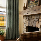 A cozy room viewpoint looking towards a rustic stone fireplace topped with a thick, rough-hewn wooden mantel and a framed landscape painting. To the left of the fireplace, a textured dark green curtain panel hangs neatly beside a window showing an autumn field, with the back of a plaid-patterned armchair in the foreground.