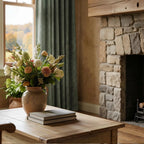 A close-up interior perspective highlighting a light wood coffee table topped with a stack of books and a ceramic vase filled with a colorful bouquet of roses and wildflowers. In the soft-focus background, a rustic stone fireplace stands to the right, and a window is decorated with textured, deep green curtains.