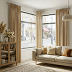 A bright, wide-angle shot of a contemporary living room with large corner windows dressed in long, light brown curtain panels. A cream-colored sofa with neutral accent pillows sits on a geometric patterned rug, flanked by a wooden glass-door cabinet on the left.