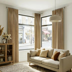A full, front-facing view of the elegant living room, showcasing the light brown curtains framing large windows that look out onto a city street. The cream sofa is styled with various warm-toned pillows, matching the geometric area rug below.