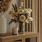 A close-up interior vignette focusing on the top of a wooden cabinet. It features a fluted white vase with dried grass, a glass vase filled with cream-colored flowers, and a scented candle, with the textured light brown curtain softly blurred in the background.