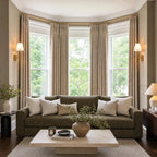 A bright, beautifully symmetrical wide shot of a living room featuring a large bay window. The window is dressed with long, light yellow-tan curtain panels and flanked by elegant brass wall sconces. A deep olive-green sofa with neutral accent pillows sits centered on a striped area rug behind a low limestone coffee table.