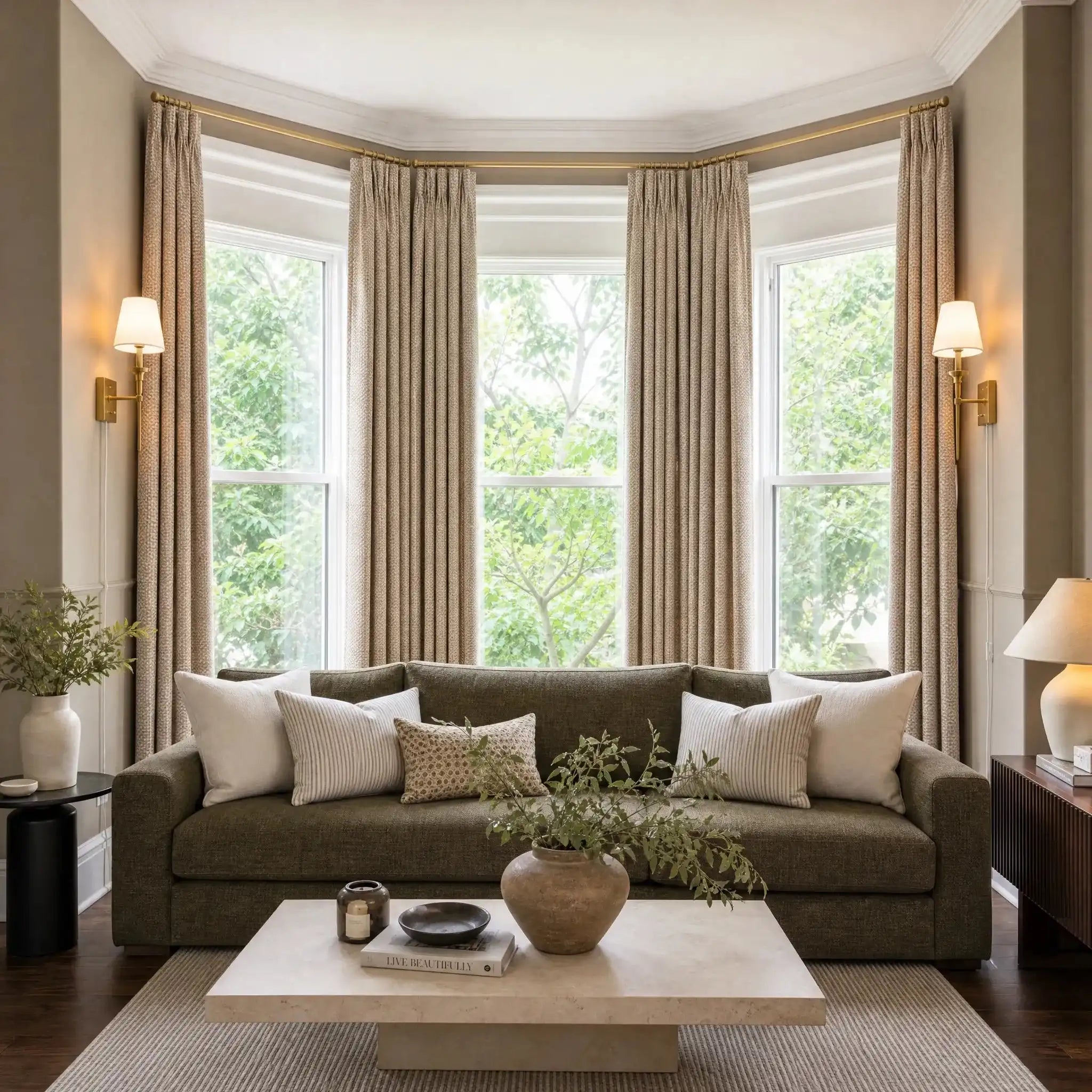 A bright, beautifully symmetrical wide shot of a living room featuring a large bay window. The window is dressed with long, light yellow-tan curtain panels and flanked by elegant brass wall sconces. A deep olive-green sofa with neutral accent pillows sits centered on a striped area rug behind a low limestone coffee table.