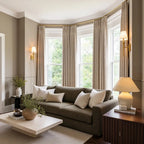 An angled full view of the elegant living room, capturing the olive-green sofa and limestone coffee table from a perspective that highlights a dark wood fluted side table with a large ceramic lamp in the right foreground. Light yellow-tan curtains frame the large bay window in the background.